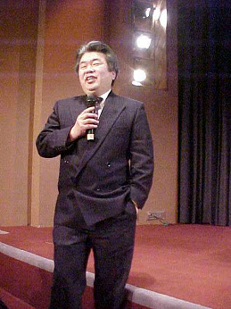 Masaki Iizuka speaking at a microphone, undated photograph