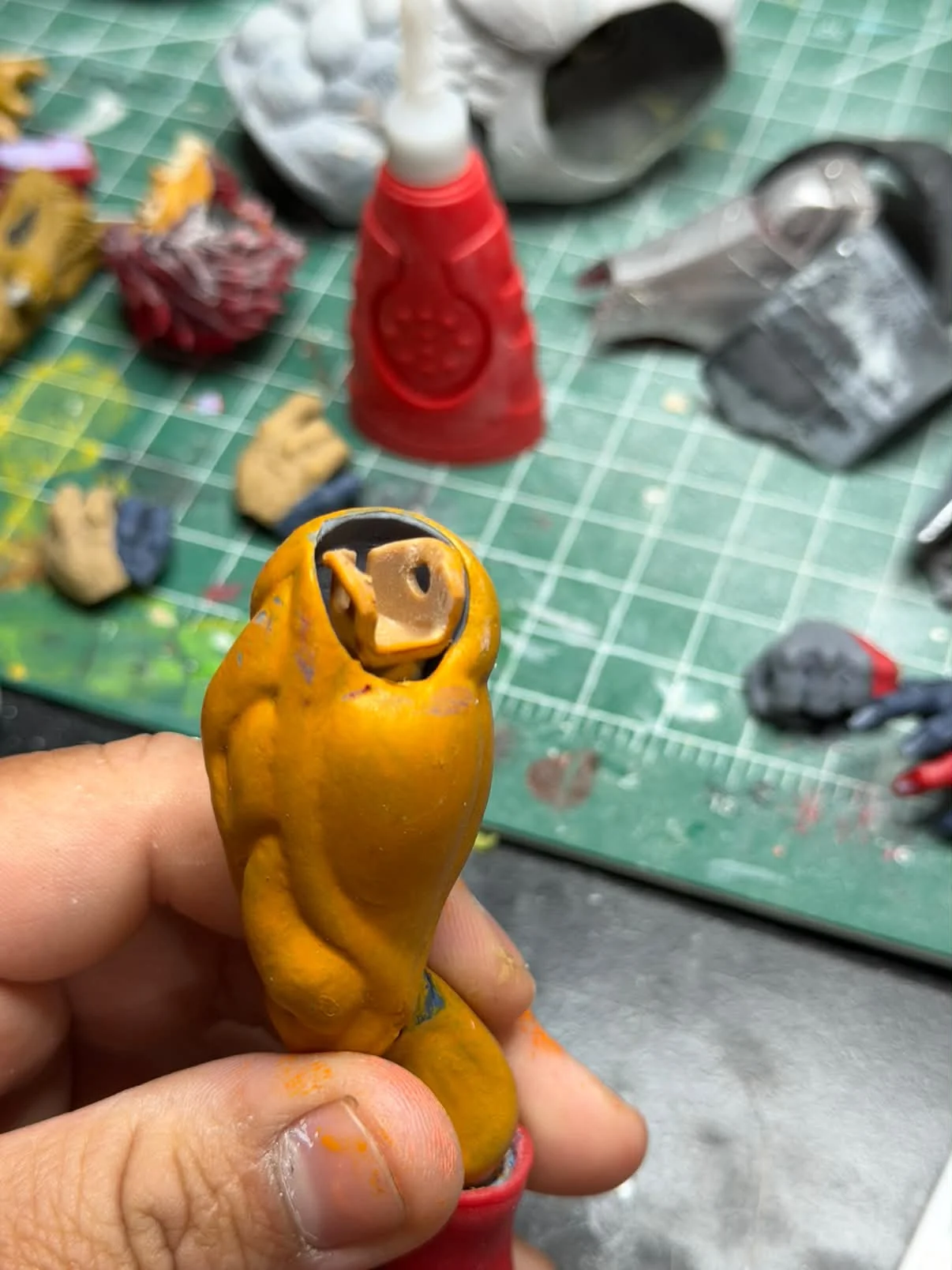 Two orange painted limb parts held between finger and thumb over a green cutting mat covered in loose figure parts