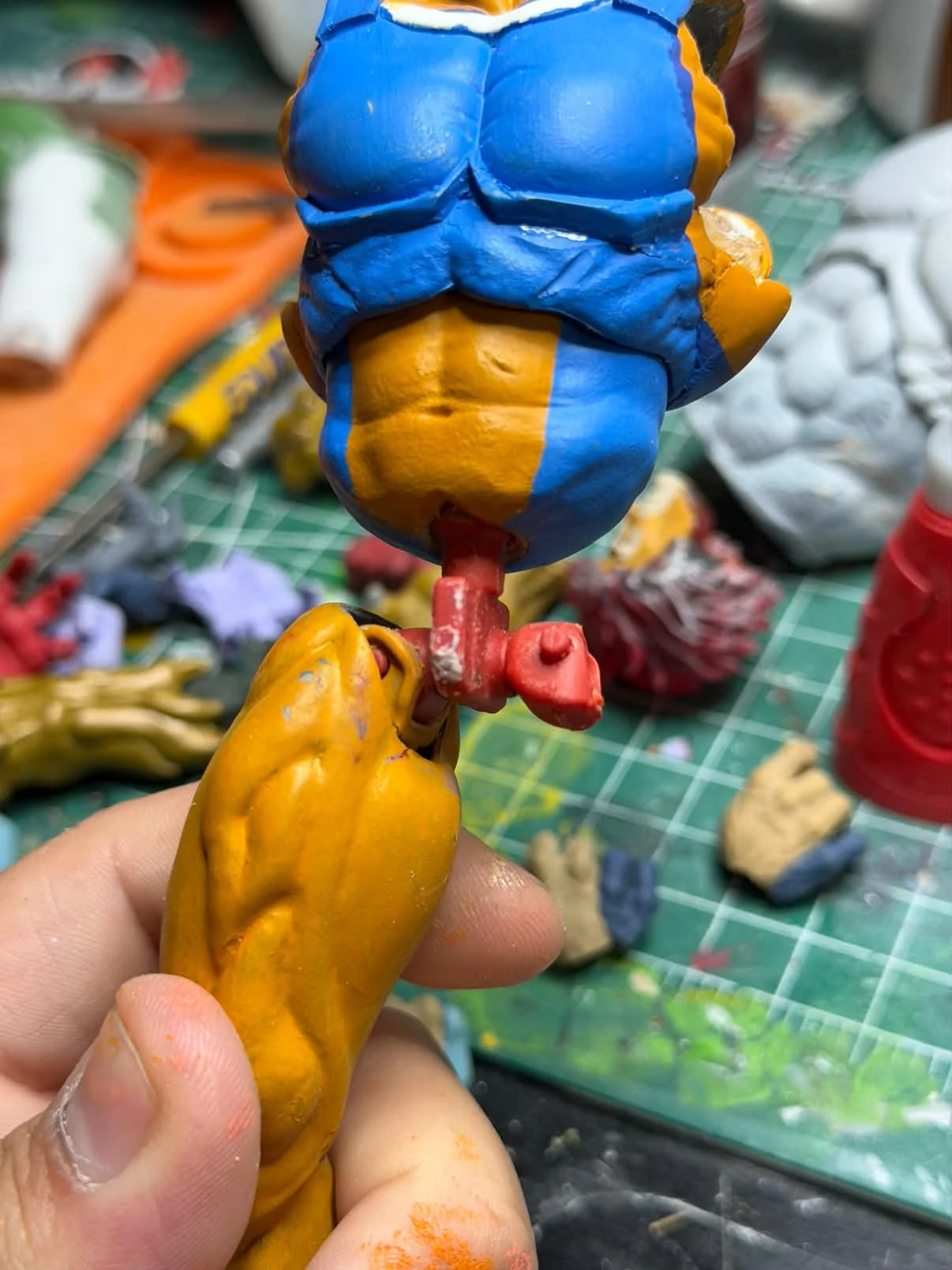 A blue and orange painted torso being test fitted against a separate orange limb part above a cluttered workbench