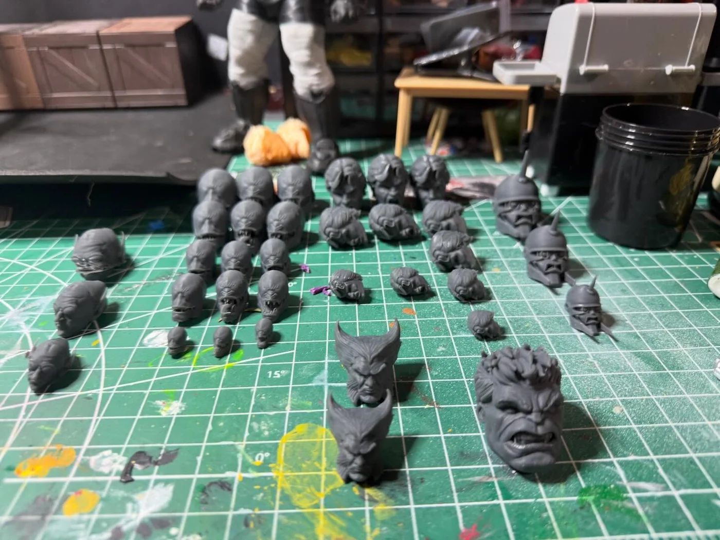 Around thirty five freshly printed grey resin heads arranged in rows on a green cutting mat, including bald skull-like heads, spiked helmets, a Wolverine style cowl and a large Hulk head at the front