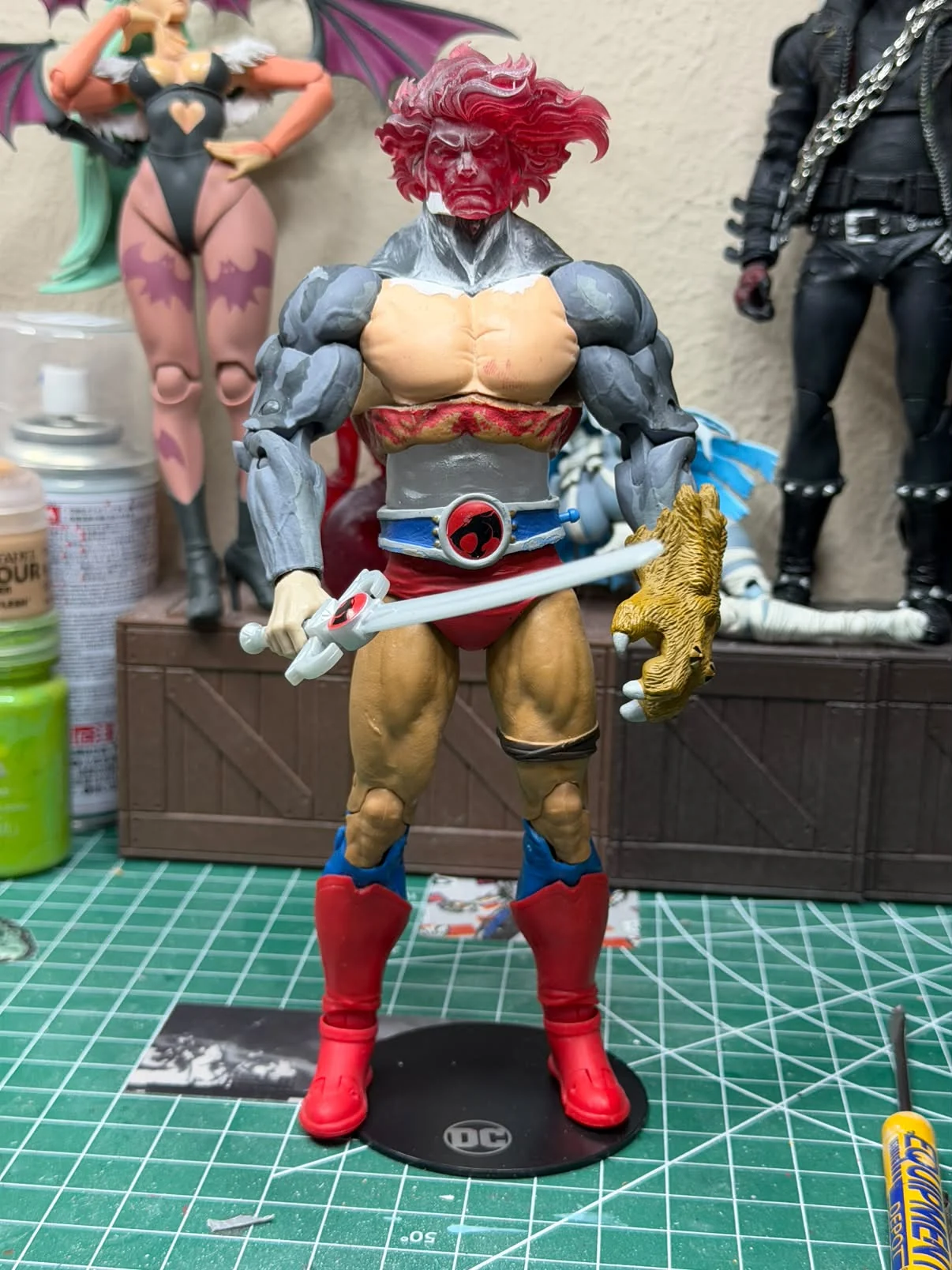Work in progress full figure standing on a display base, red 3D printed head, grey unpainted arms, red trunks, holding an unpainted Sword of Omens and a claw shield