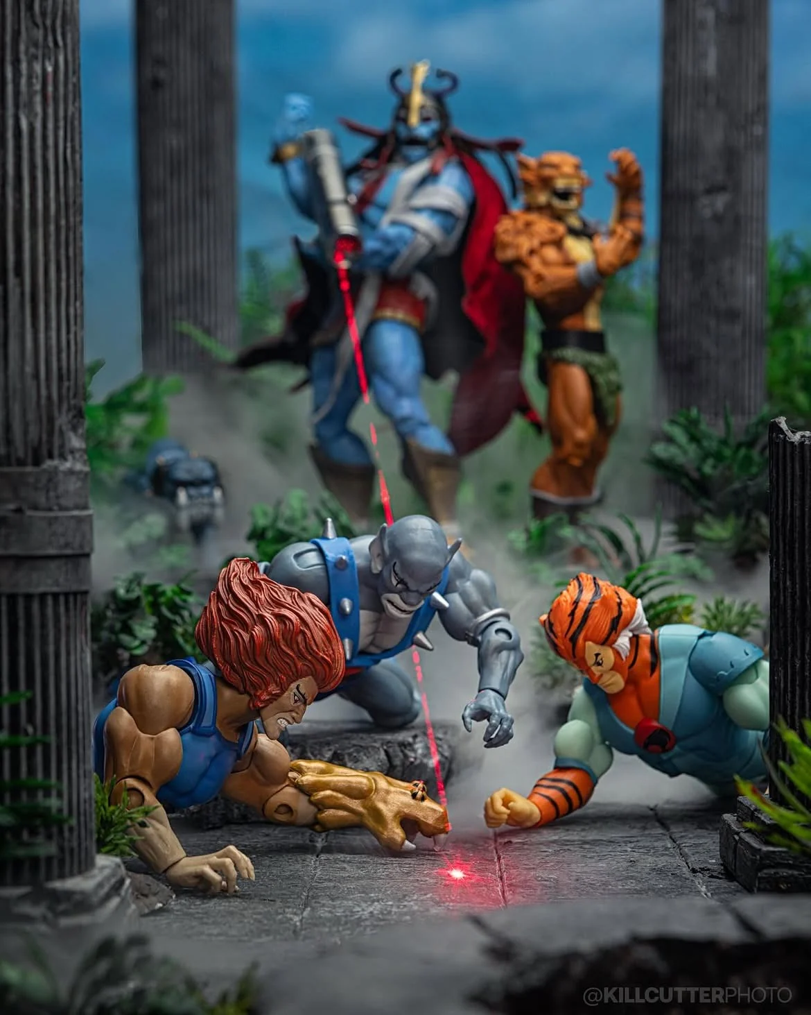 Lion-O, Panthro and Tygra scramble on a stone floor chasing a red laser dot while Mumm-Ra and Jackalman watch from a ruined temple behind them