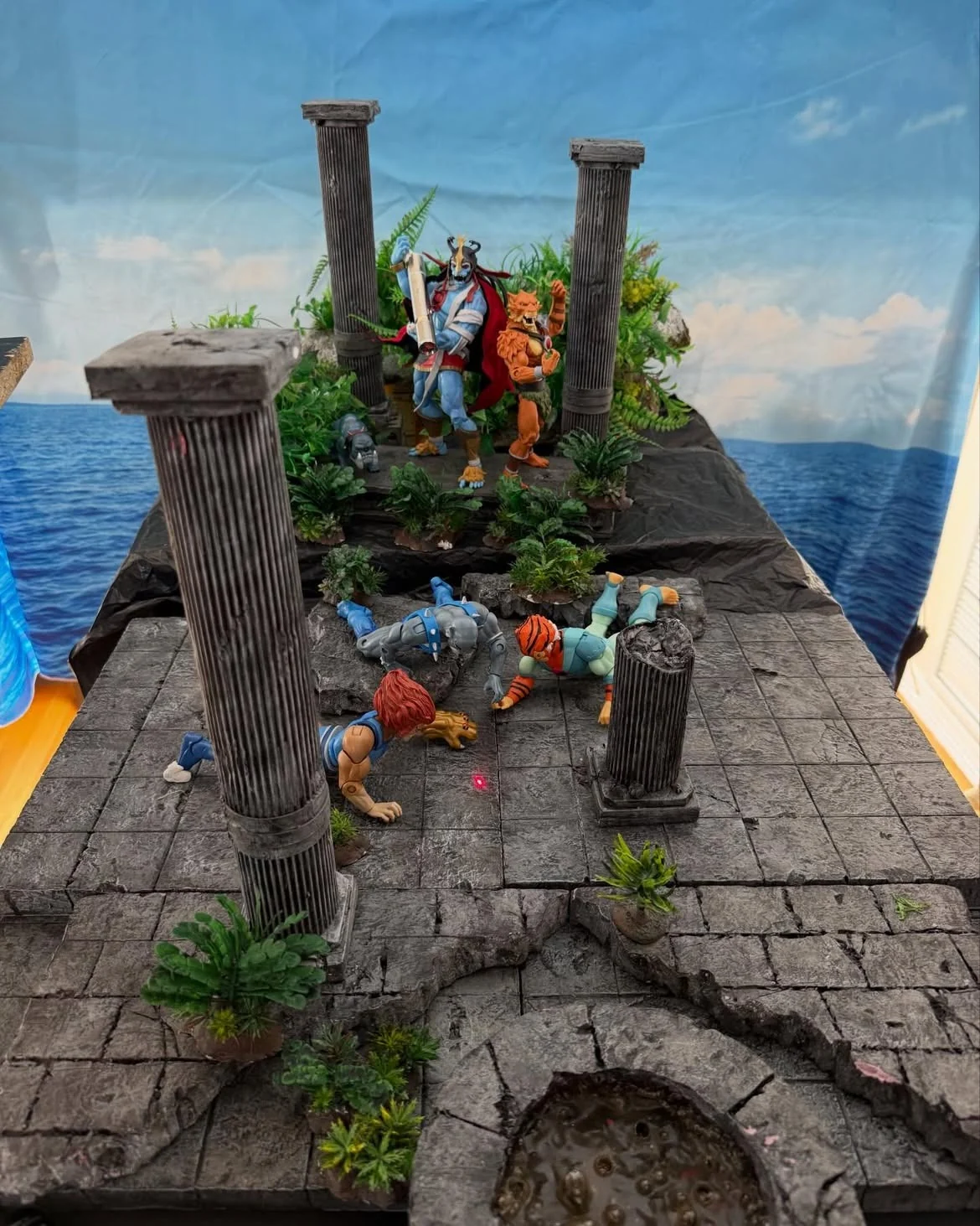 The same temple scene photographed from above in ordinary room light, showing the diorama sitting on a table in front of a printed sea and sky backdrop