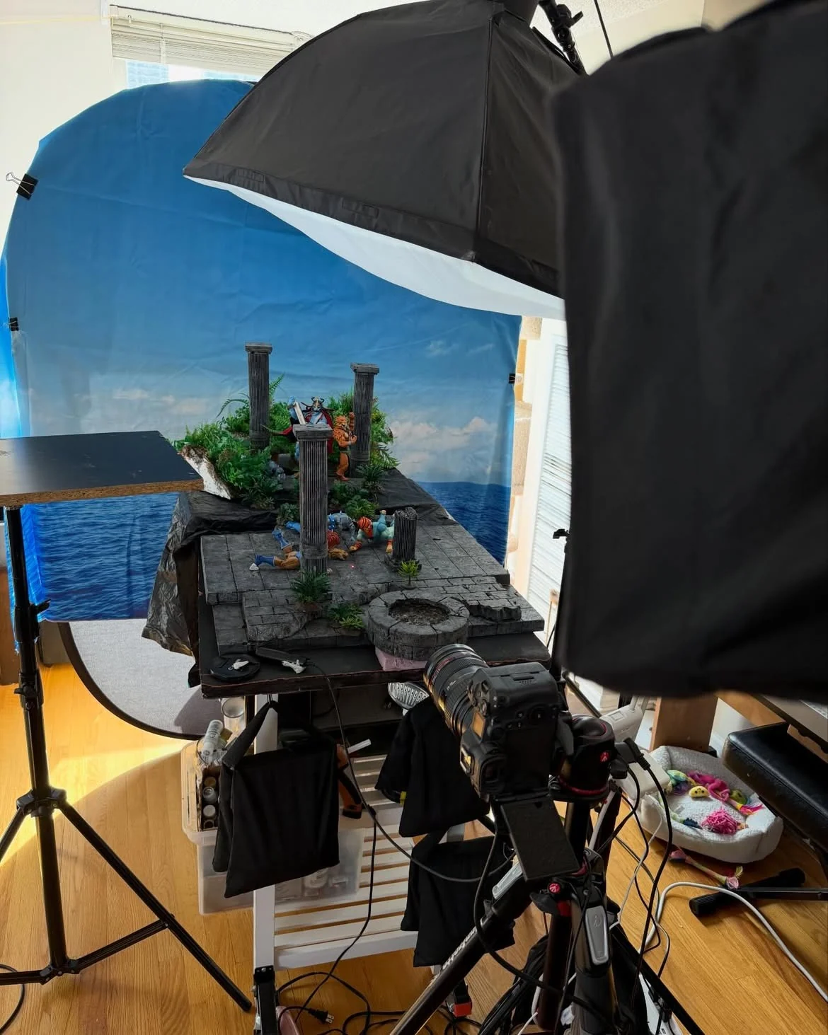Behind the scenes view of the shoot: the diorama on a rolling cart in a living room, lit by two softboxes with a camera on a tripod in the foreground