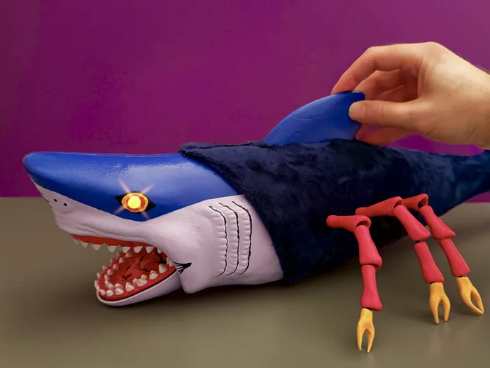 A hand lifting the dorsal fin of the custom Black Widow Shark, showing the plush body and the lit eye